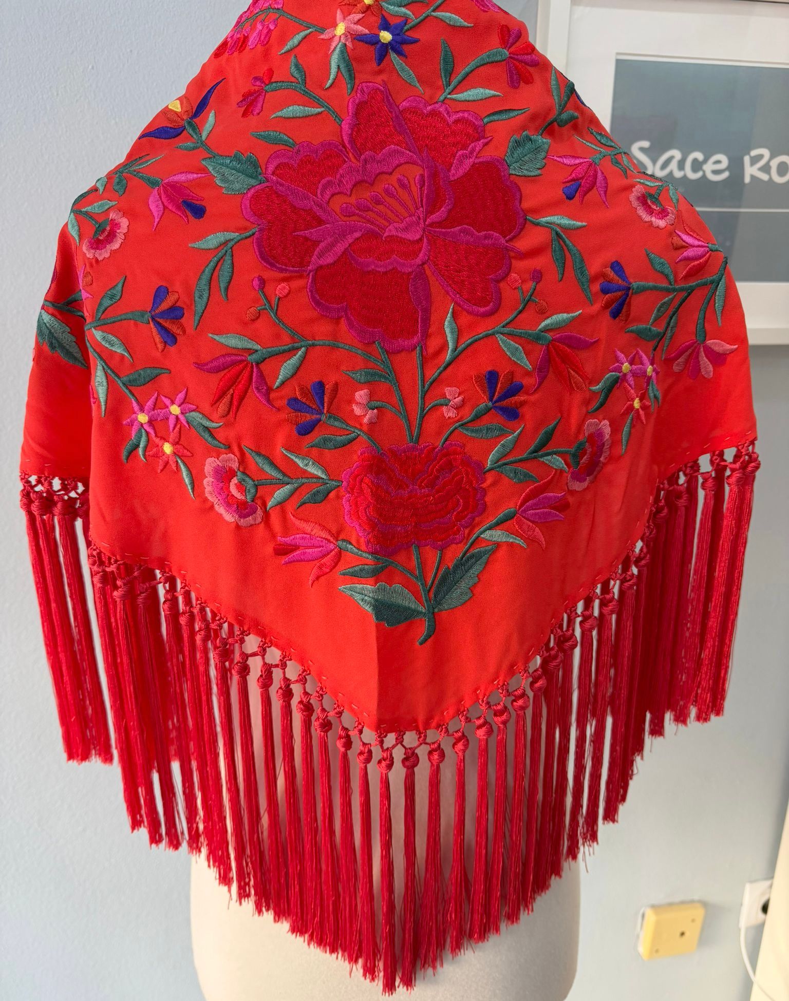 Mantón triangular, de crepe bordado de flores en colores vivos con flecos por la parte de abajo, con un enrejado que termina con un borlón y el fleco es de 15 cm.